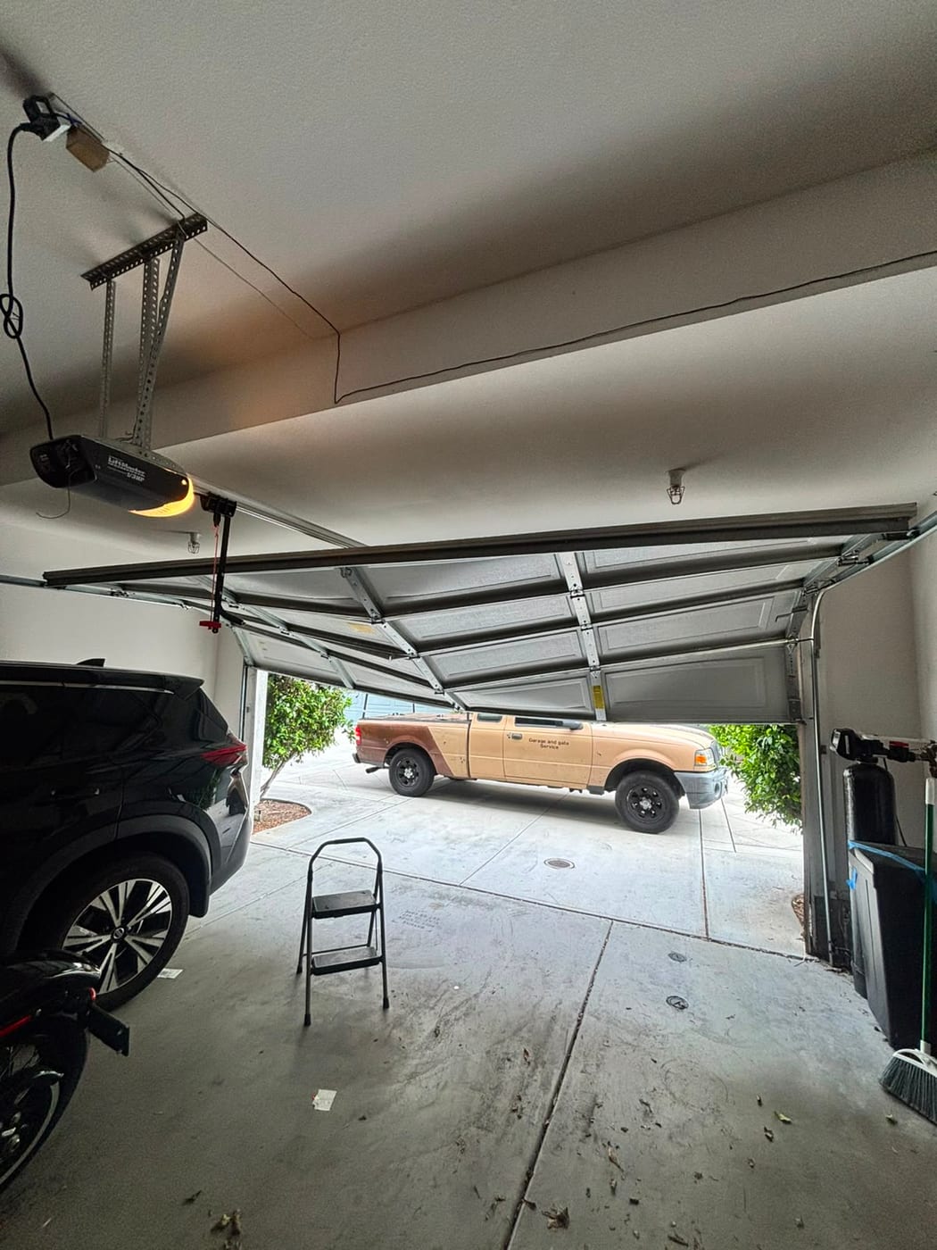 A residential garage door part way open with the opener running, and the Garage and Gate Palace service truck parked on the driveway outside