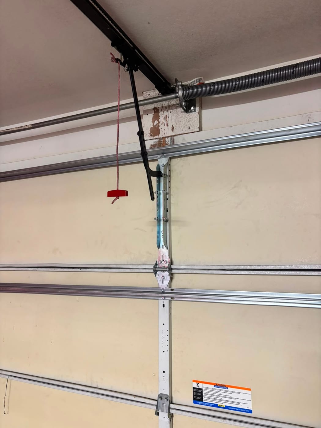 Torsion spring and bar mounted above a garage door, with the opener rail, door arm and red emergency release cord visible